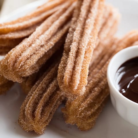 Freshly fried churros are stacked on parchment paper, steam rising from the warm cinnamon-sugar pastries ready to be dipped.
